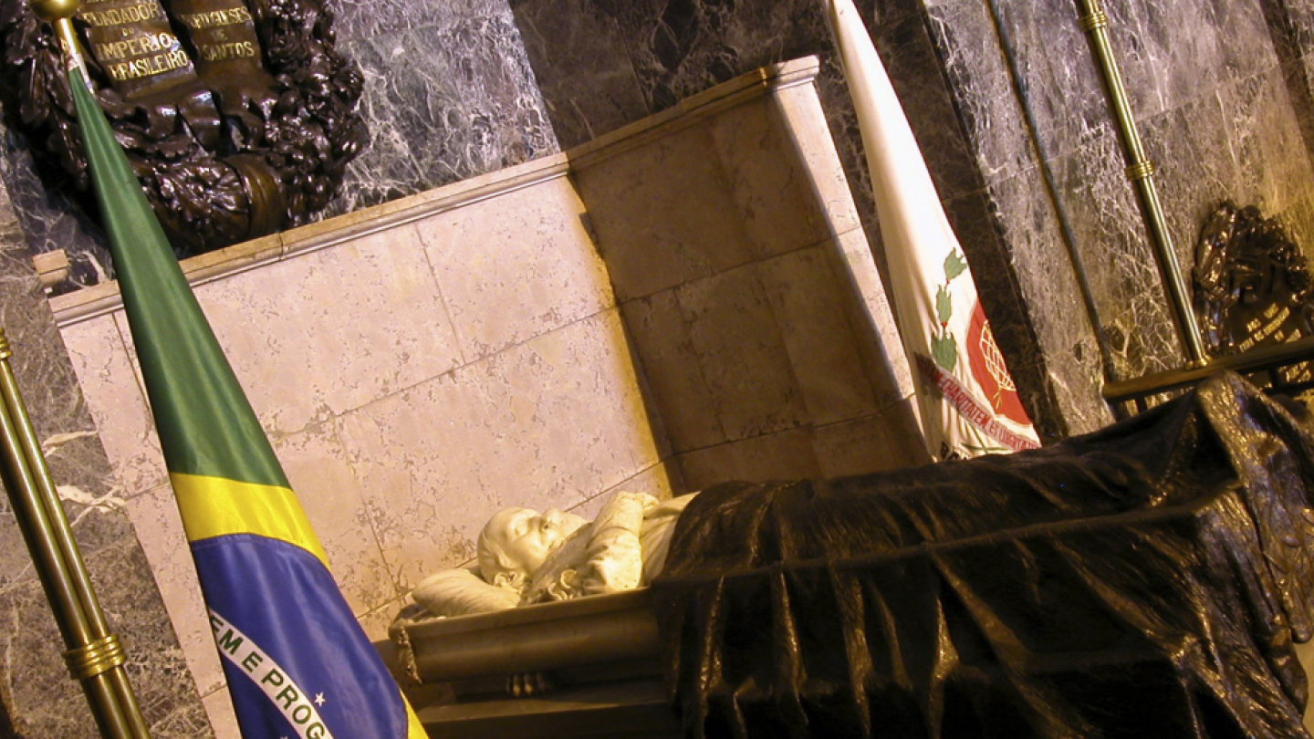 Uma fotografia em plano inclinado captura um sarcófago ou tumba monumental parcialmente coberta com tecido preto, ladeada por bandeiras (incluindo a do Brasil em primeiro plano) dentro de um ambiente interno de mármore escuro