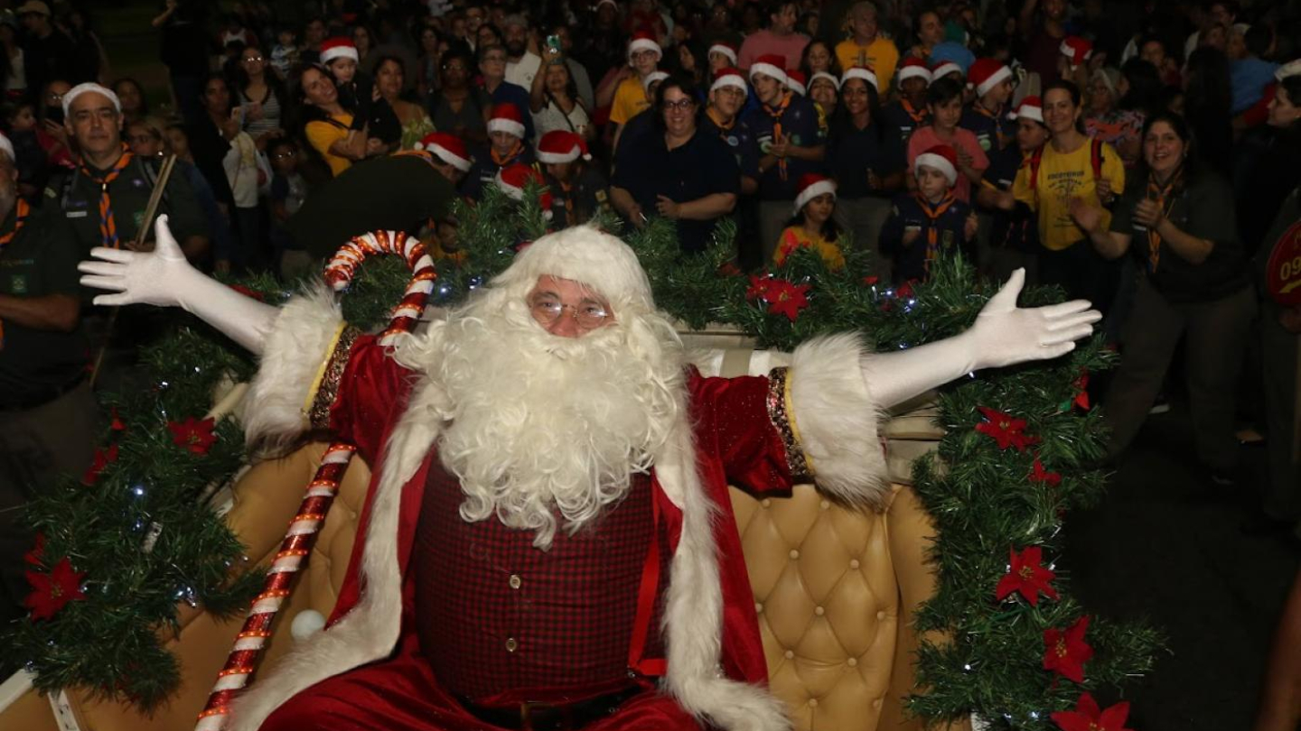 Em um momento festivo noturno, Papai Noel está sentado com os braços abertos, em um trenó ornamentado com guirlandas e flores de Natal e, ao fundo, uma multidão de pessoas observam usando gorros e roupas natalinas