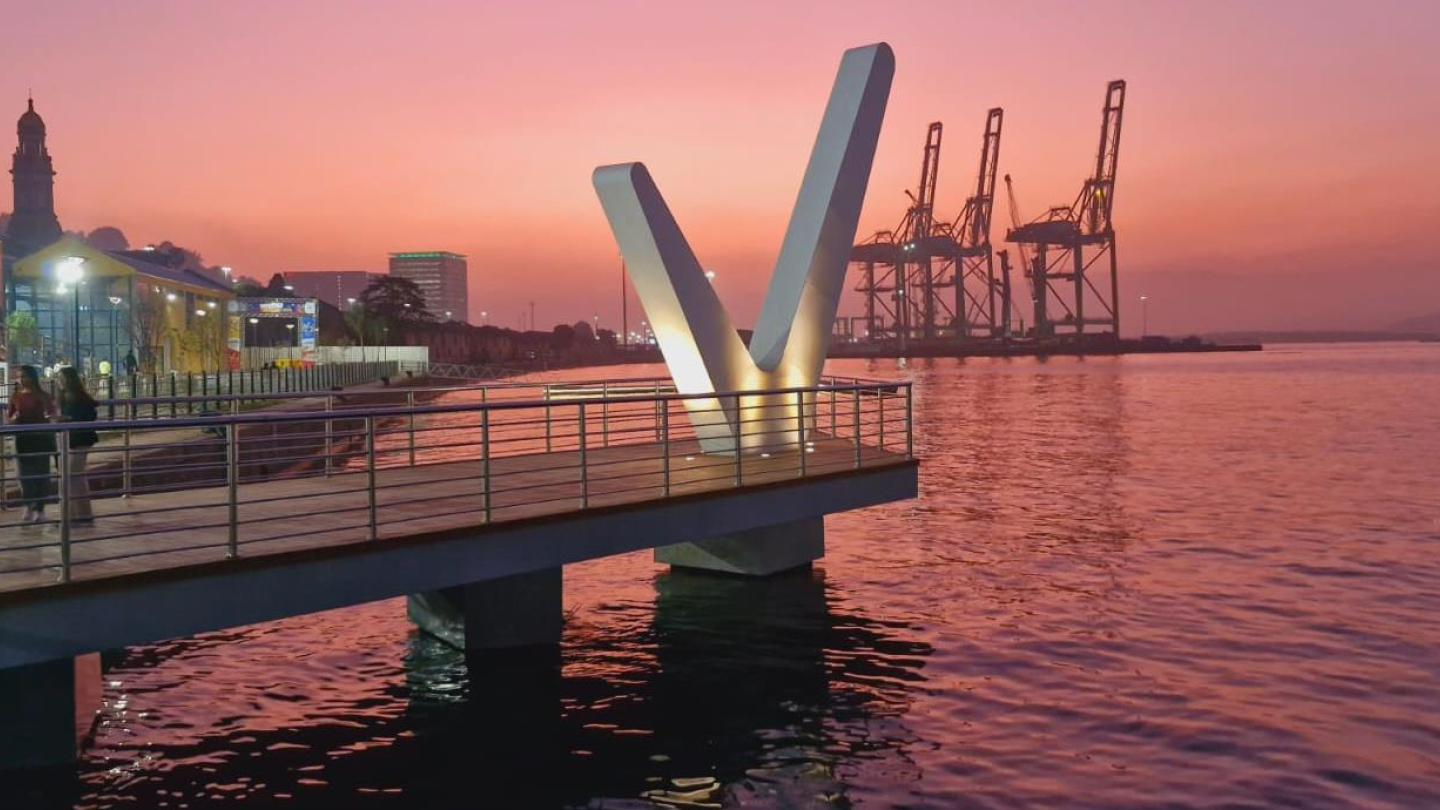 Pôr do sol rosado no Parque Valongo, em Santos, com deck iluminado, escultura em “V”, água refletindo as cores do céu e guindastes do porto ao fundo