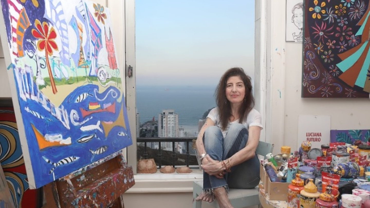 Mulher sentada em um ateliê, ao lado de telas em cores vibrantes e diversos potes e latas de tintas