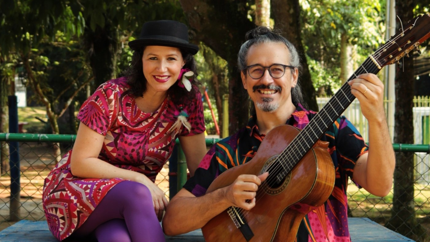 Um casal sorridente e colorido (uma mulher de vestido magenta, meias roxas e chapéu, e um homem de óculos e camisa estampada, segurando um violão) posa em um banco azul contra um fundo de árvores em um parque