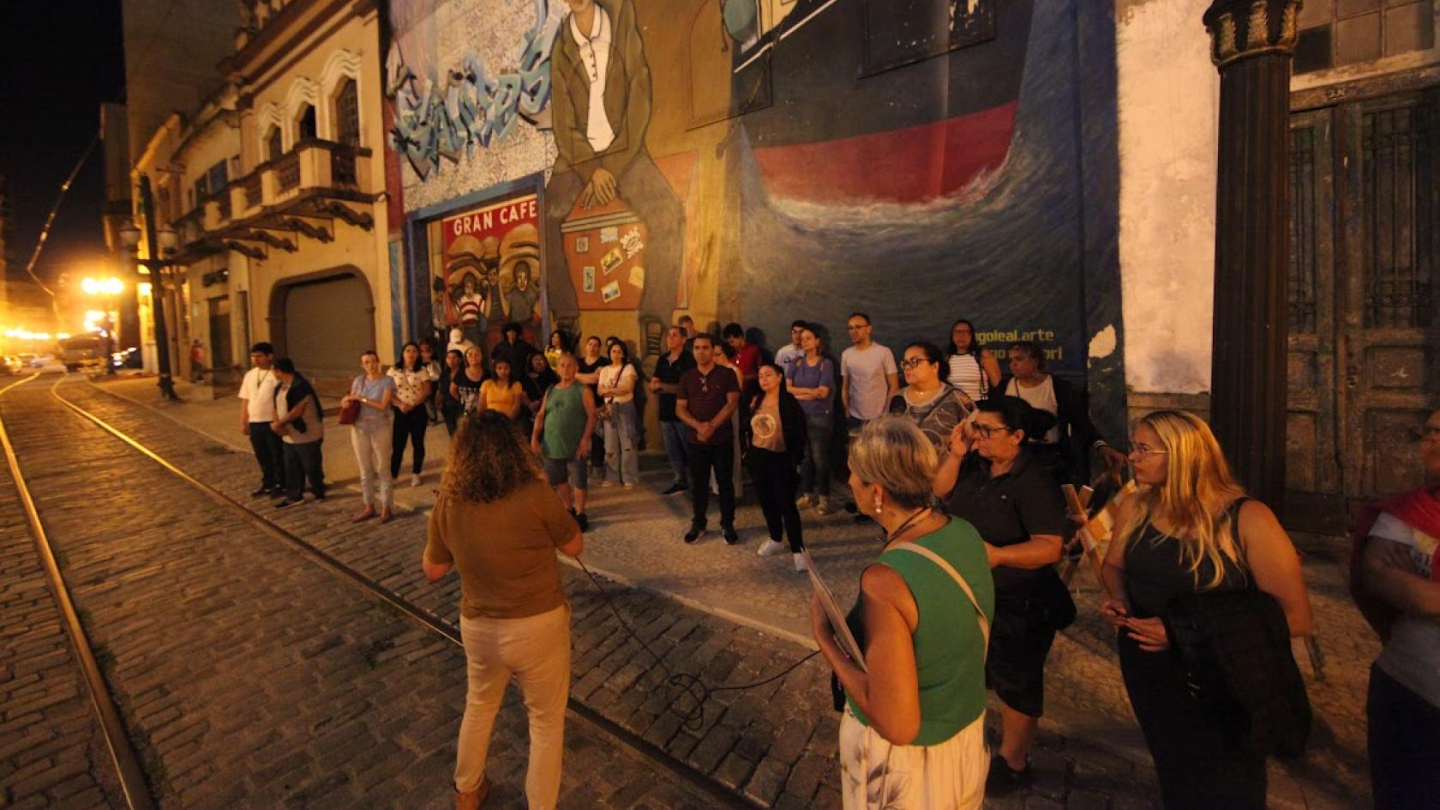 Um grupo de pessoas participa de uma visita guiada noturna em uma rua de paralelepípedos com trilhos de bonde, em frente a um grande mural artístico em uma construção antiga