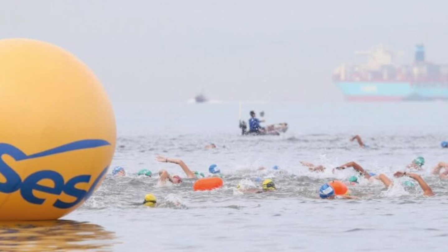 Uma grande boia amarela com o logotipo do Sesc está em primeiro plano, enquanto nadadores competem em mar aberto, com um navio cargueiro desfocado ao longe e um bote de resgate no fundo