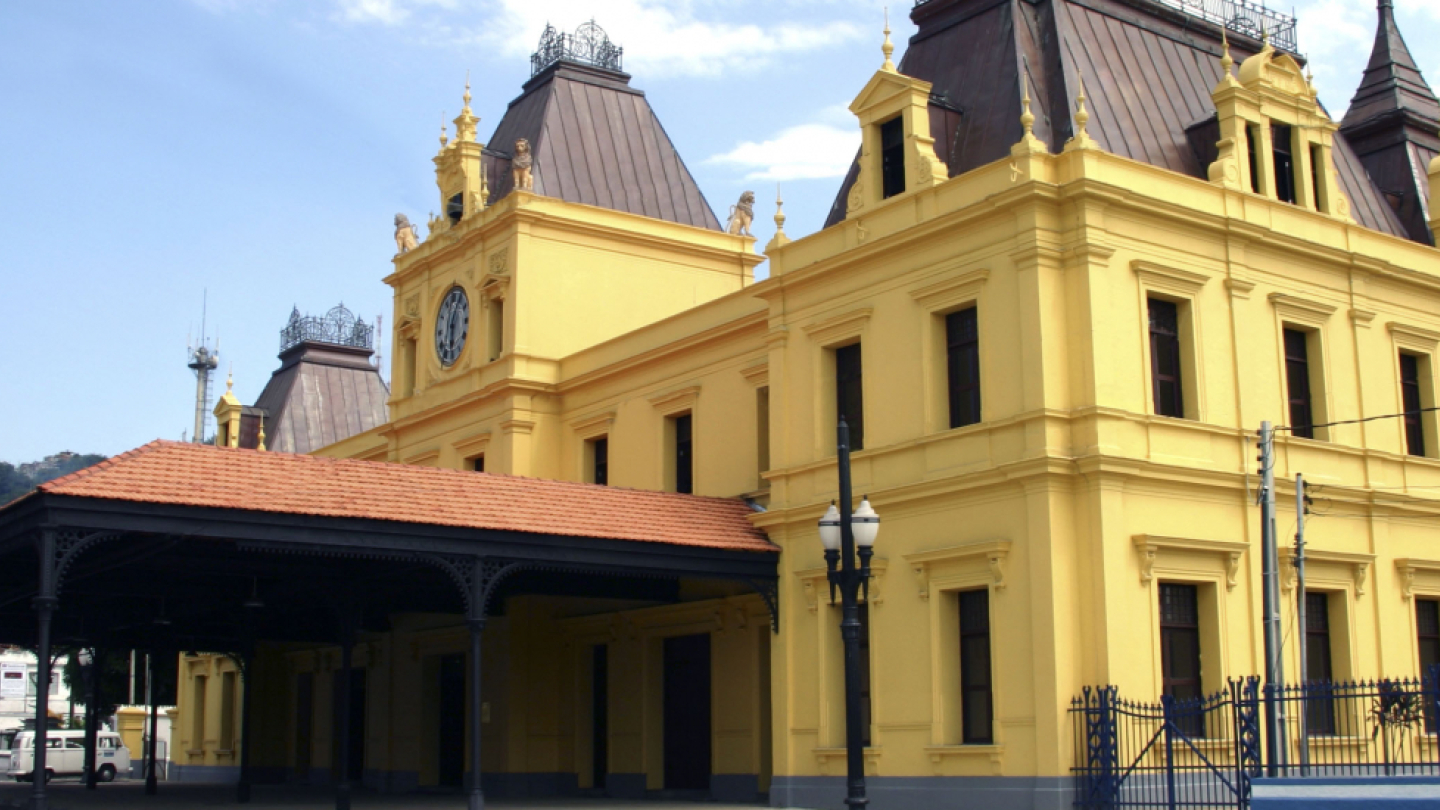 Prédio histórico de fachada amarela com detalhes arquitetônicos clássicos e telhados inclinados em tom marrom. No centro, há uma torre com relógio. À frente, uma cobertura metálica preta e postes de iluminação completam a cena. O céu está claro e há morros verdes ao fundo.