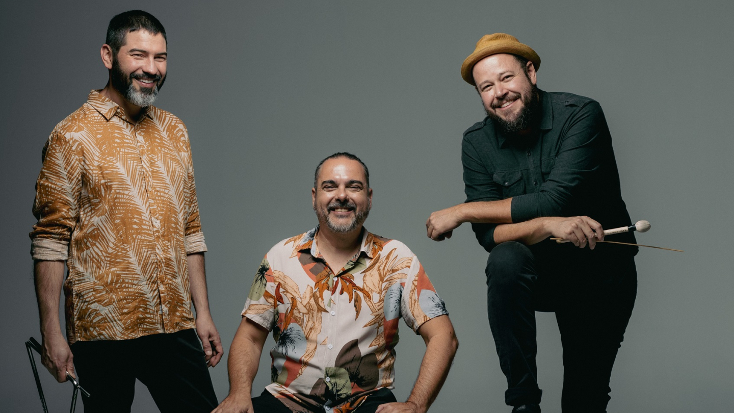 Imagem de três músicos brasileiros sorridentes, sendo um com um triângulo, outro sentado em um caixote com uma sanfona à sua frente, e o terceiro com um bumbo e baquetas