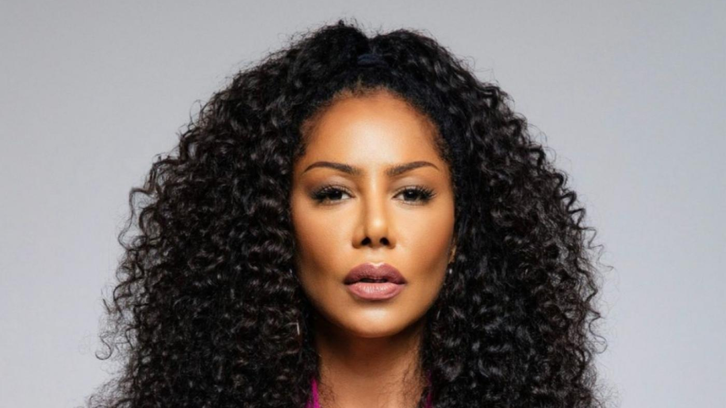 Mulher com longos e volumosos cabelos cacheados, vestindo uma peça de roupa magenta com decote em V profundo e posando com uma expressão séria em um fundo cinza claro