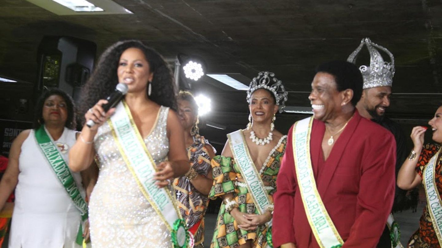 Valéria Valenssa, com um microfone na mão e faixa de Embaixadora do Carnaval, sorri ao lado de Neguinho da Beija-Flor, também Embaixador do Carnaval, e da Corte Carnavalesca de Santos
