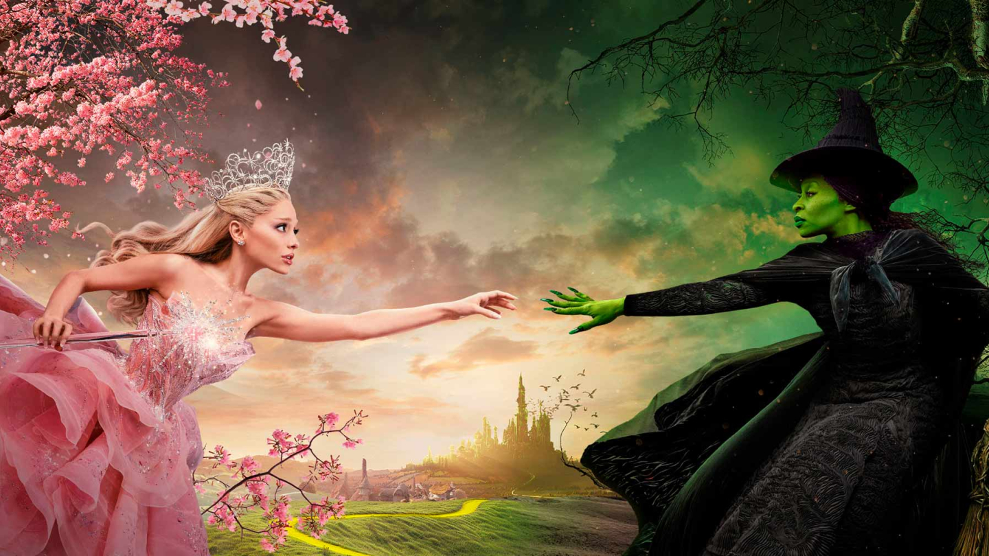 Uma imagem promocional vibrante e dramática mostra duas mulheres estendendo as mãos uma para a outra: uma, a bruxa Glinda (loira, em rosa, sob uma árvore florida) e a outra, a bruxa Elphaba (verde, em preto, sob uma árvore seca), com a estrada de tijolos amarelos e um castelo ao fundo, representando o universo de Wicked