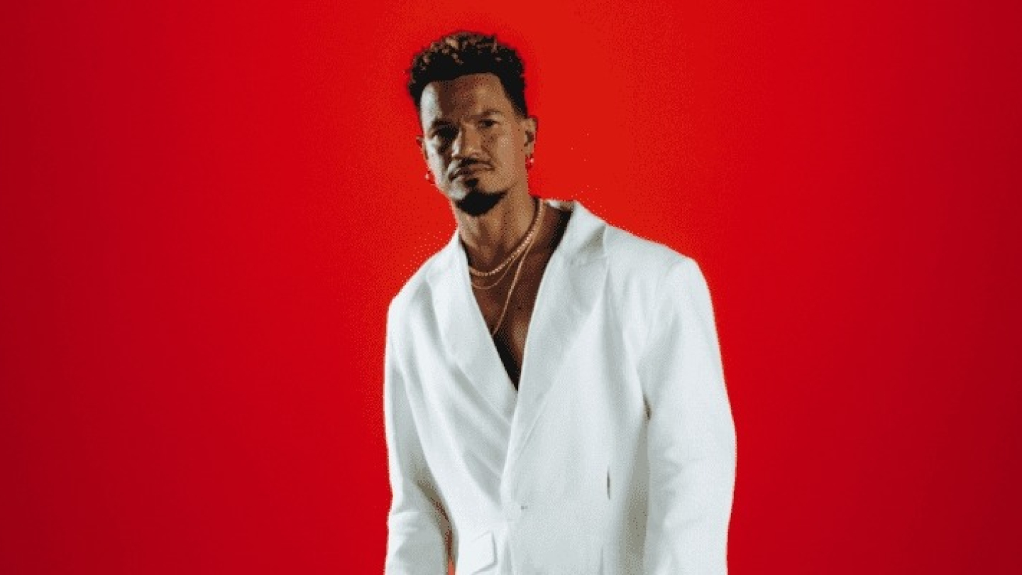 Um homem veste um elegante blazer branco aberto com colares dourados e posa com uma expressão séria em frente a um vibrante fundo vermelho