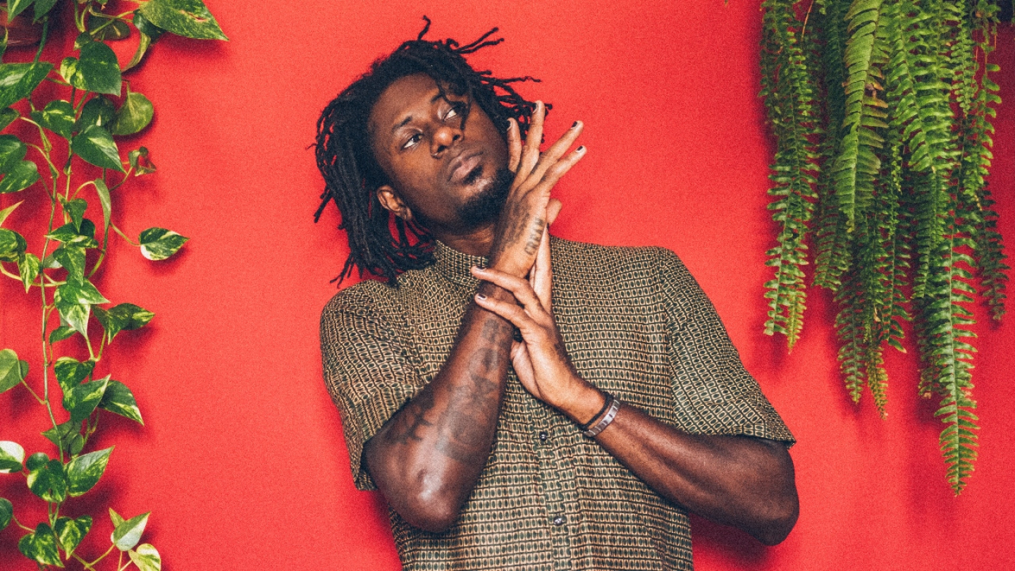 O rapper brasileiro Zudizilla, de dreadlocks e camisa estampada, está em frente a um fundo vermelho vibrante, com plantas verdes em ambos os lados, e as suas mãos sobrepostas à frente do corpo