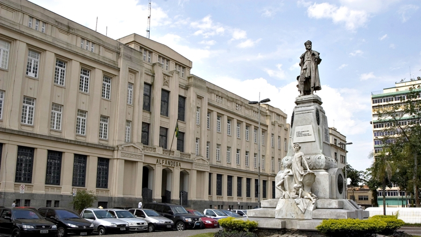 A imagem mostra uma praça urbana com um grande prédio histórico ao fundo e um monumento escultórico em primeiro plano.