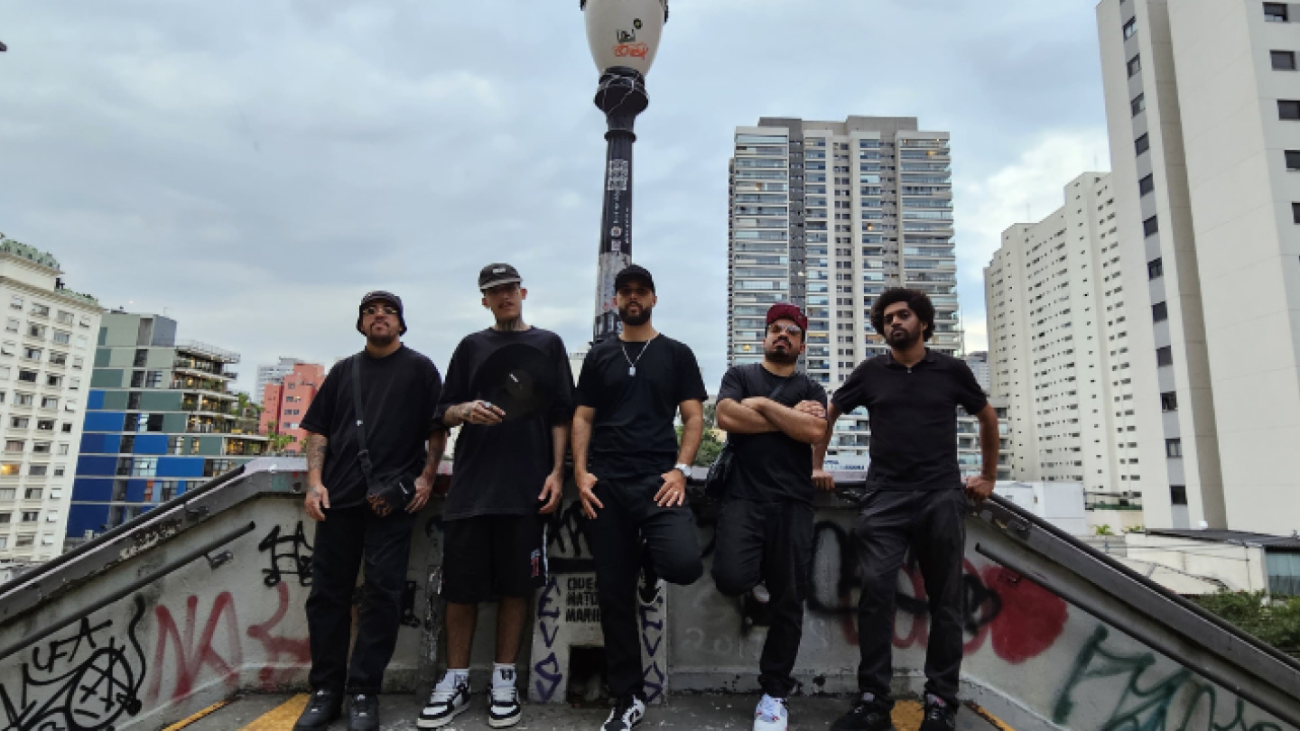 Cinco homens, todos vestidos de preto e com bonés/toucas, posam em uma área coberta de grafites sob um poste de luz, tendo como fundo o céu nublado e a paisagem urbana com prédios altos