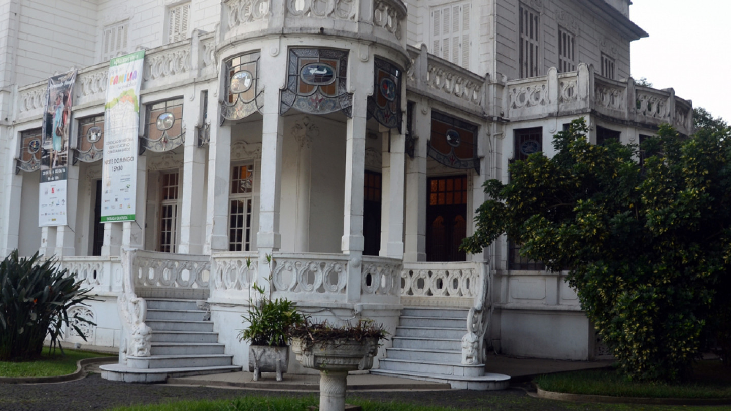 A imagem mostra um elegante edifício histórico de cor clara, com forte inspiração eclética e art nouveau, cercado por jardins bem cuidados. A construção se destaca pela fachada ornamentada, com sacadas curvas, colunas esguias e delicados elementos decorativos em relevo.