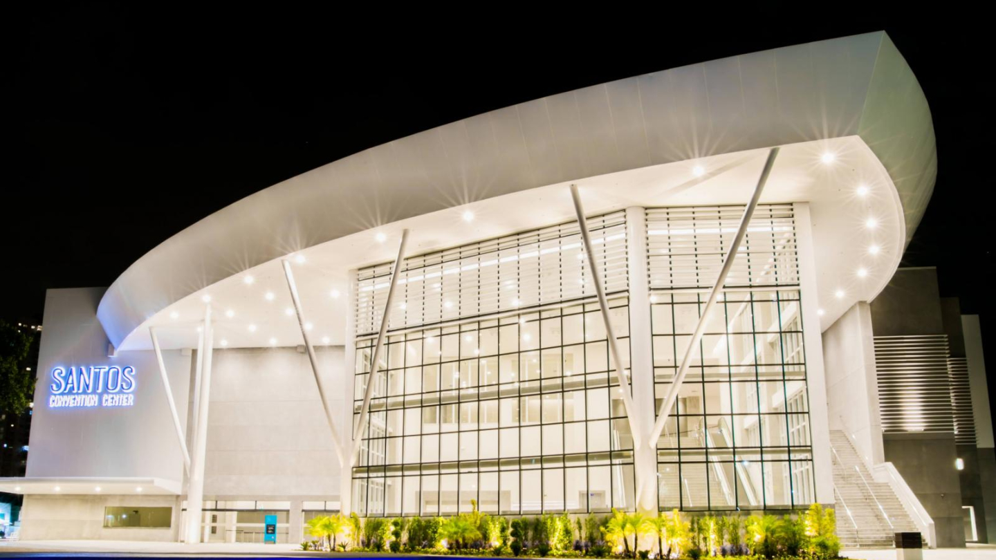 Imagem noturna do Santos Convention Center, prédio moderno de fachada envidraçada e estrutura branca com colunas inclinadas. A iluminação destaca o formato curvo do telhado, com jardins e espelho d’água à frente.