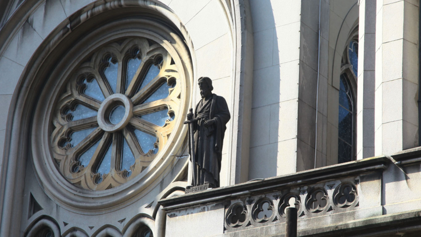 A imagem mostra um recorte da fachada de uma igreja de grande porte, com forte inspiração neogótica. O destaque principal é o grande vitral circular em forma de rosácea, com divisões radiais e detalhes ornamentais em pedra, típico desse estilo arquitetônico. A fachada é clara, em tons de bege e cinza, com linhas verticais marcantes e arcos altos que reforçam a sensação de imponência.
