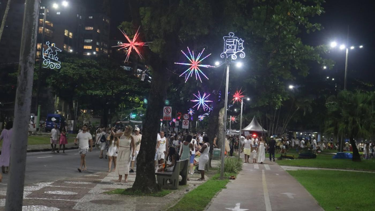 A imagem mostra uma cena noturna movimentada à beira da praia, provavelmente durante uma celebração ou evento de fim de ano. A avenida e o calçadão estão repletos de pessoas caminhando, conversando, tirando fotos e aproveitando a noite — muitas delas vestidas de branco, o que reforça o clima típico da virada do ano.