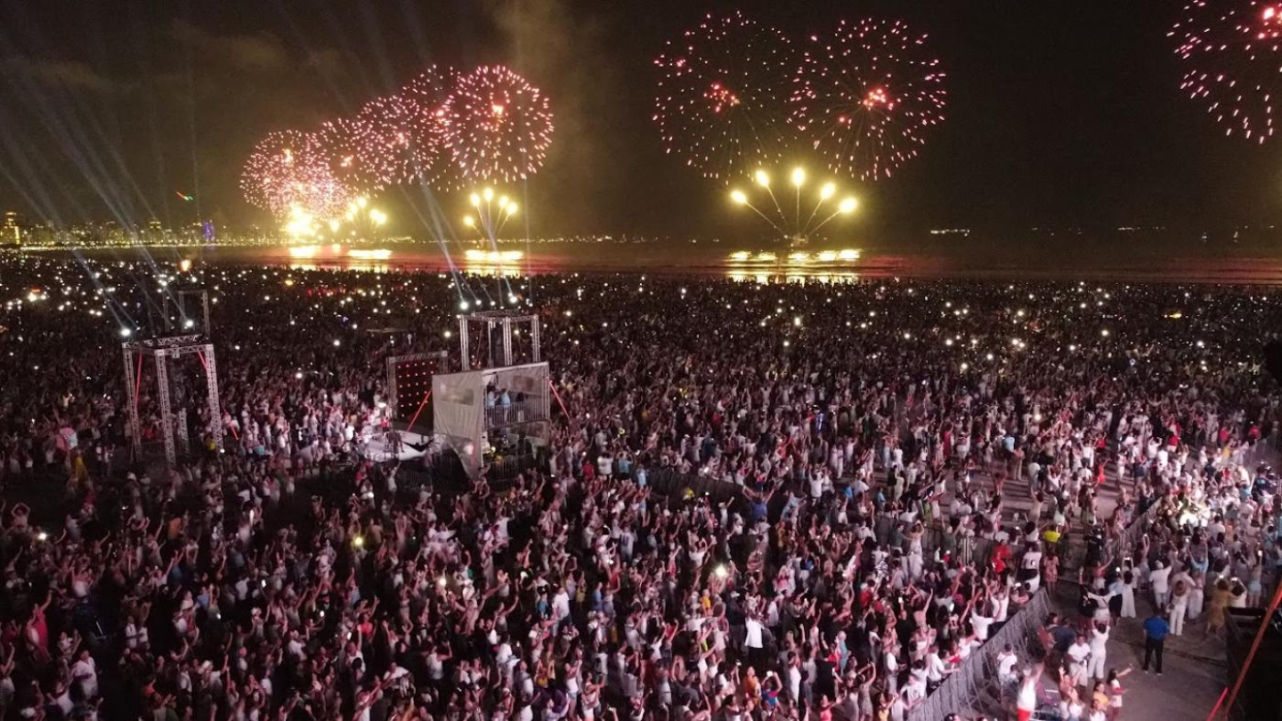 Imagem noturna de uma grande multidão reunida na orla da praia. Milhares de pessoas ocupam a areia, muitas com braços erguidos e celulares iluminados. Ao fundo, sobre o mar, fogos de artifício coloridos explodem no céu, refletindo na água e iluminando a paisagem urbana à esquerda.
