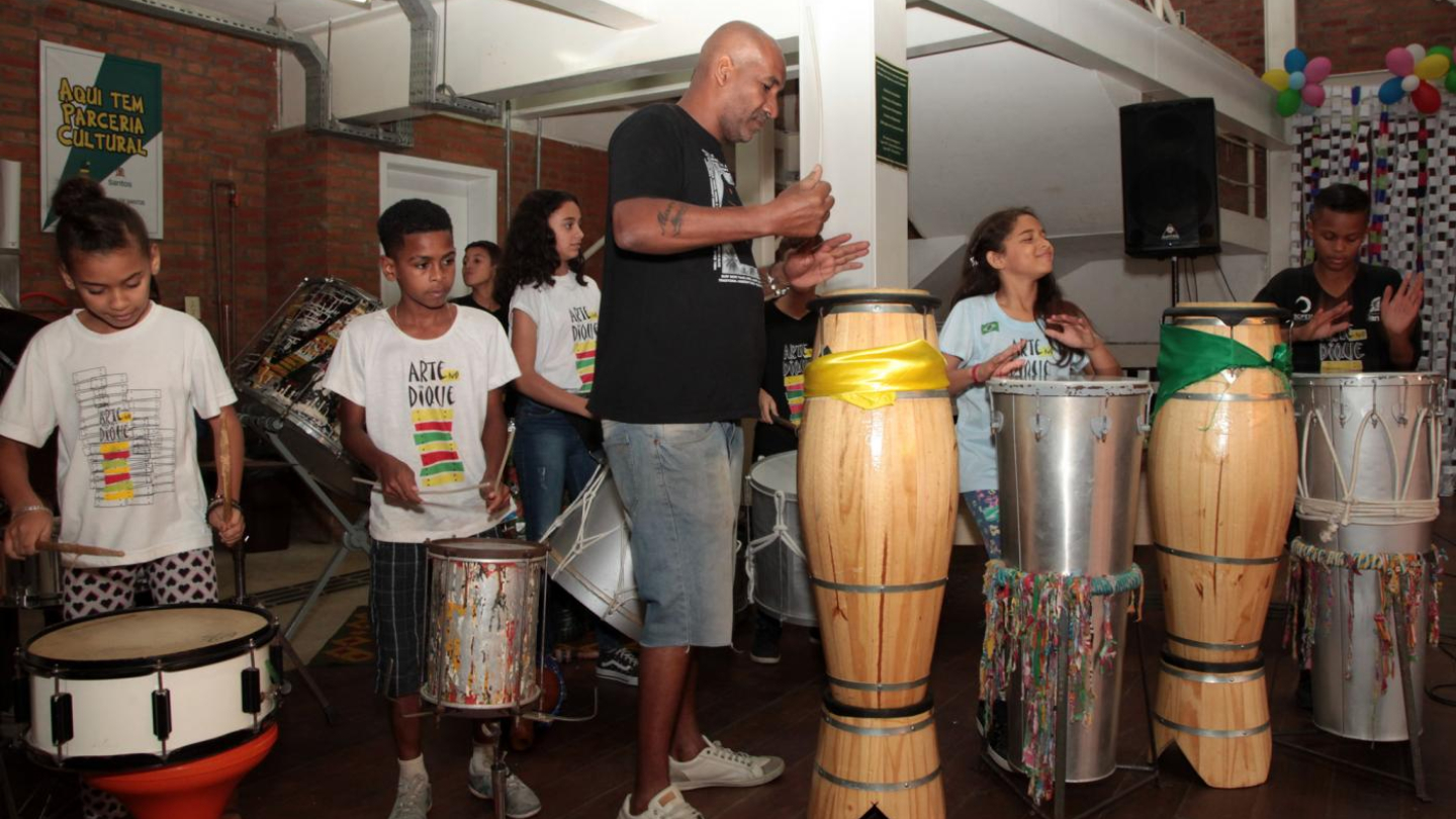  A fotografia registra um momento de oficina ou apresentação de percussão, onde um instrutor adulto e várias crianças, algumas vestindo camisetas "Arte Dique", tocam diversos tipos de tambores em um espaço cultural interno