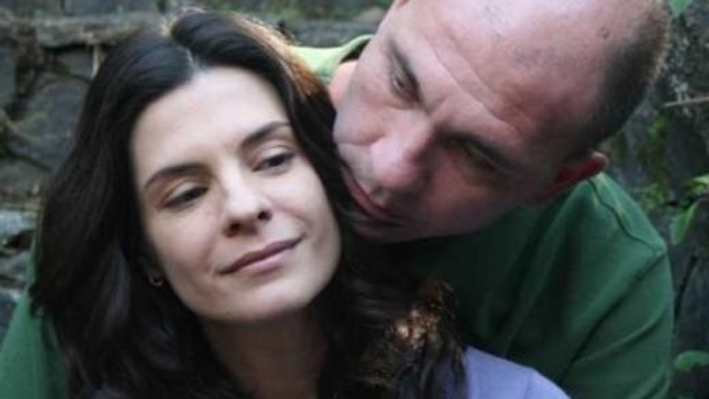 A imagem é um close-up de um casal em um ambiente externo, mostrando uma mulher sorrindo levemente e um homem careca com camisa verde inclinando a cabeça sobre o ombro dela