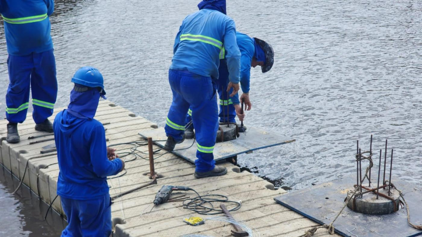 A foto mostra três trabalhadores vestindo uniformes de cor azul e equipamentos de proteção, incluindo capacetes e botas. Eles estão sobre uma plataforma flutuante instalada na água, possivelmente para algum tipo de obra hidráulica ou manutenção.