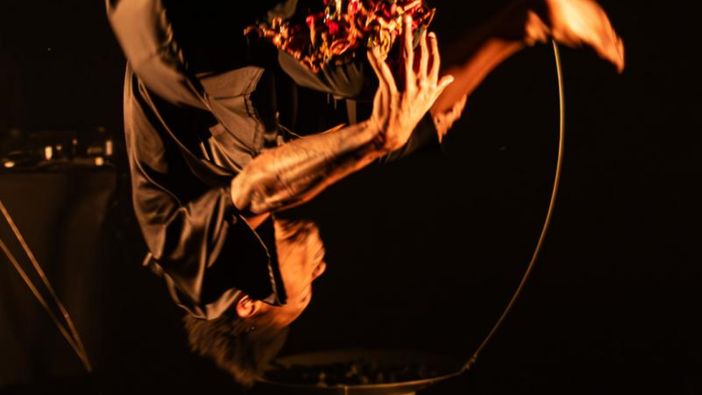 A imagem é uma fotografia artística de palco, em fundo escuro, que captura um bailarino em movimento de cambalhota ou salto acrobático com o corpo curvado e os pés para cima