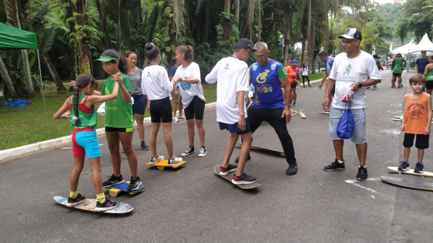 A imagem mostra várias pessoas, incluindo adultos e crianças, praticando manobras em skates e pranchas com rodinhas, em um evento ao ar livre em um parque.