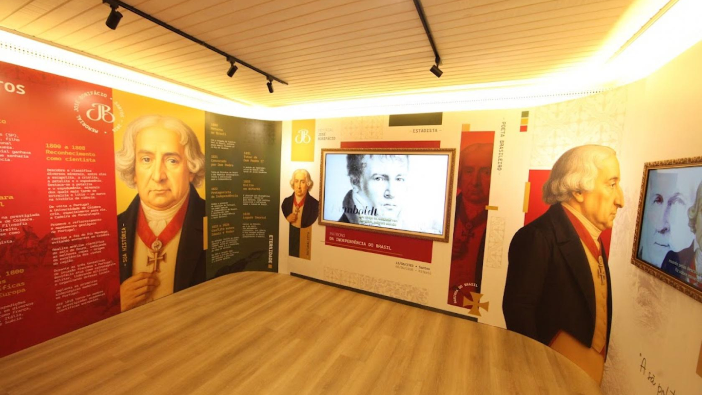 A imagem mostra uma sala de exposição moderna, bem iluminada, dedicada à vida e trajetória de José Bonifácio de Andrada e Silva. As paredes são totalmente envelopadas por painéis coloridos — em tons de vermelho, verde, amarelo e branco — que apresentam textos informativos, datas importantes e títulos que contam diferentes fases de sua atuação, como estadista, cientista e patriota da Independência do Brasil.