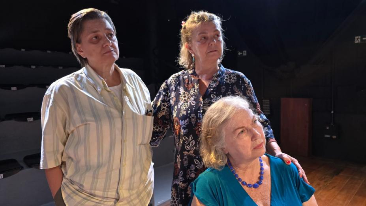 A imagem mostra três mulheres, em um palco escuro com assentos de teatro visíveis ao fundo, onde tanto a mulher sentada à frente quanto as outras duas (em pé) olham para cima