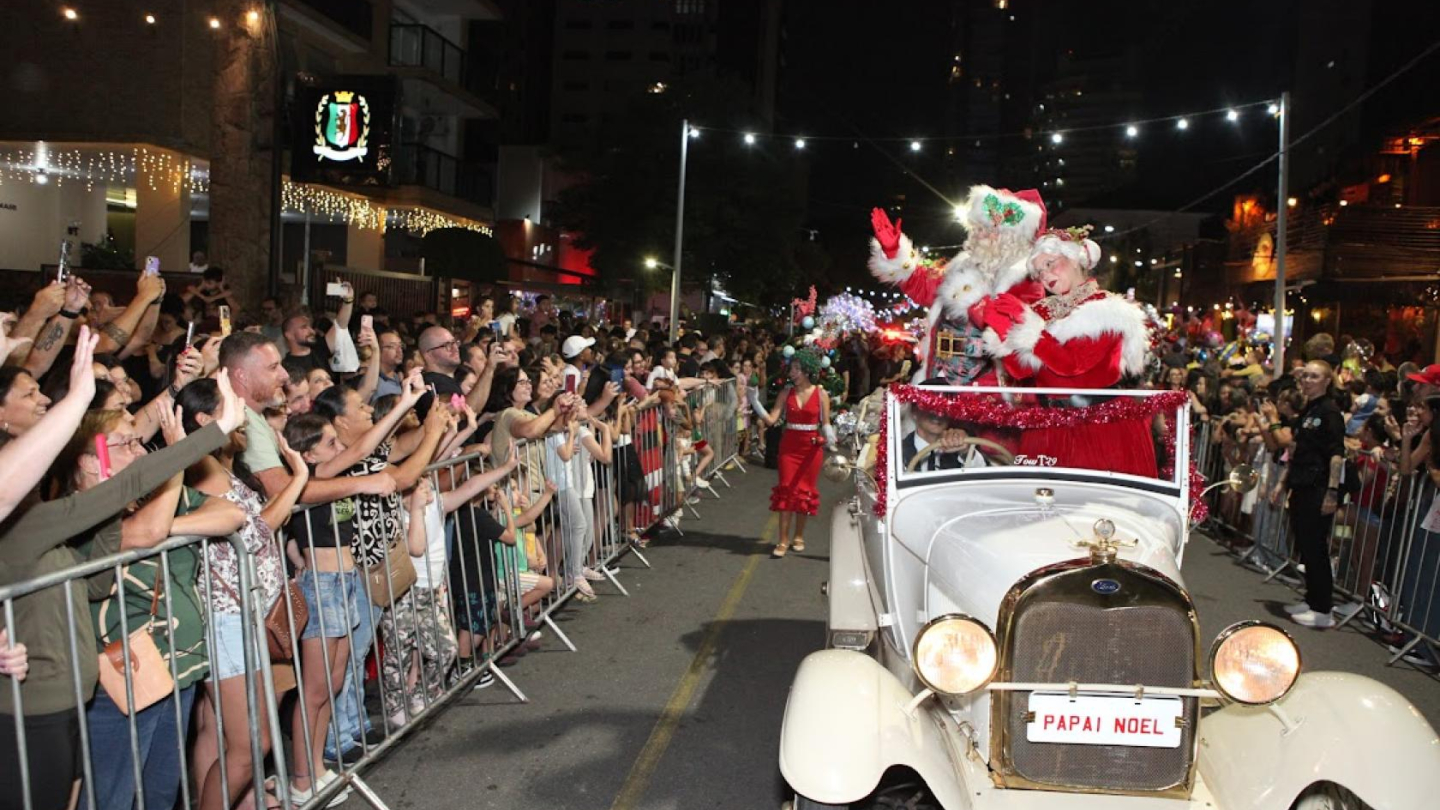 Papai e Mamãe Noel em um carro branco, aberto, como um desfile, com centenas de pessoas observando e tirando fotos.