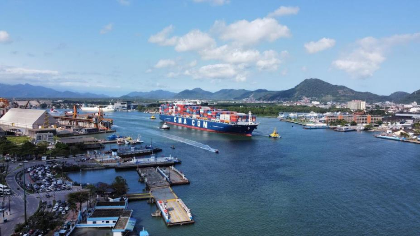 O ChatGPT disse:  A imagem mostra uma vista aérea belíssima do Porto de Santos em um dia claro e ensolarado. No centro da cena, navega um enorme navio porta-contêineres da companhia CMA CGM, carregado com dezenas de contêineres coloridos empilhados. Ele é acompanhado por rebocadores que o auxiliam na manobra pelo estuário.