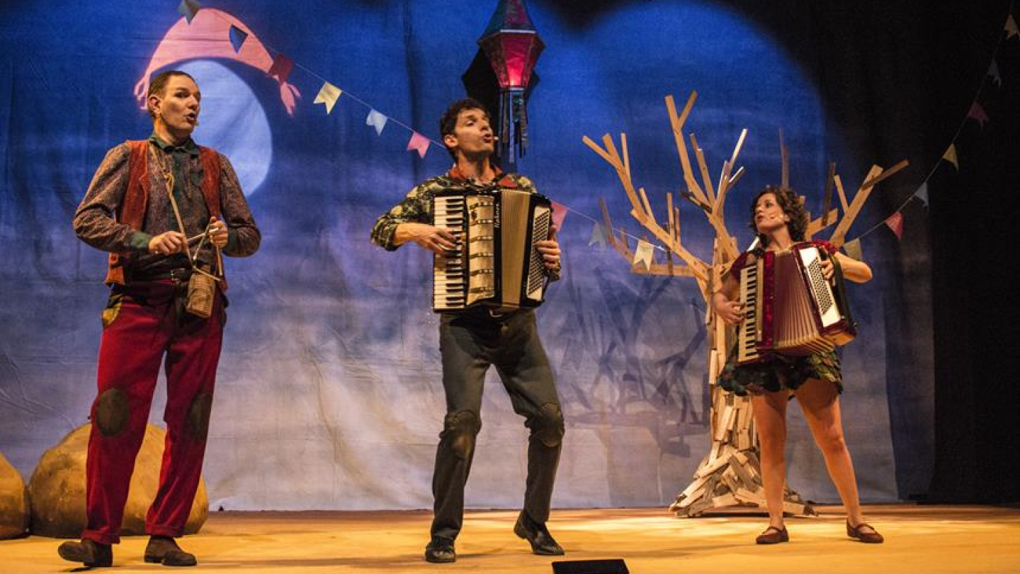 Três artistas se apresentam em um palco de teatro com cenário noturno (lua cheia e bandeirinhas) e uma árvore seca, sendo que dois deles (um homem e uma mulher) tocam acordeão e o terceiro (homem) toca um instrumento de percussão pequeno