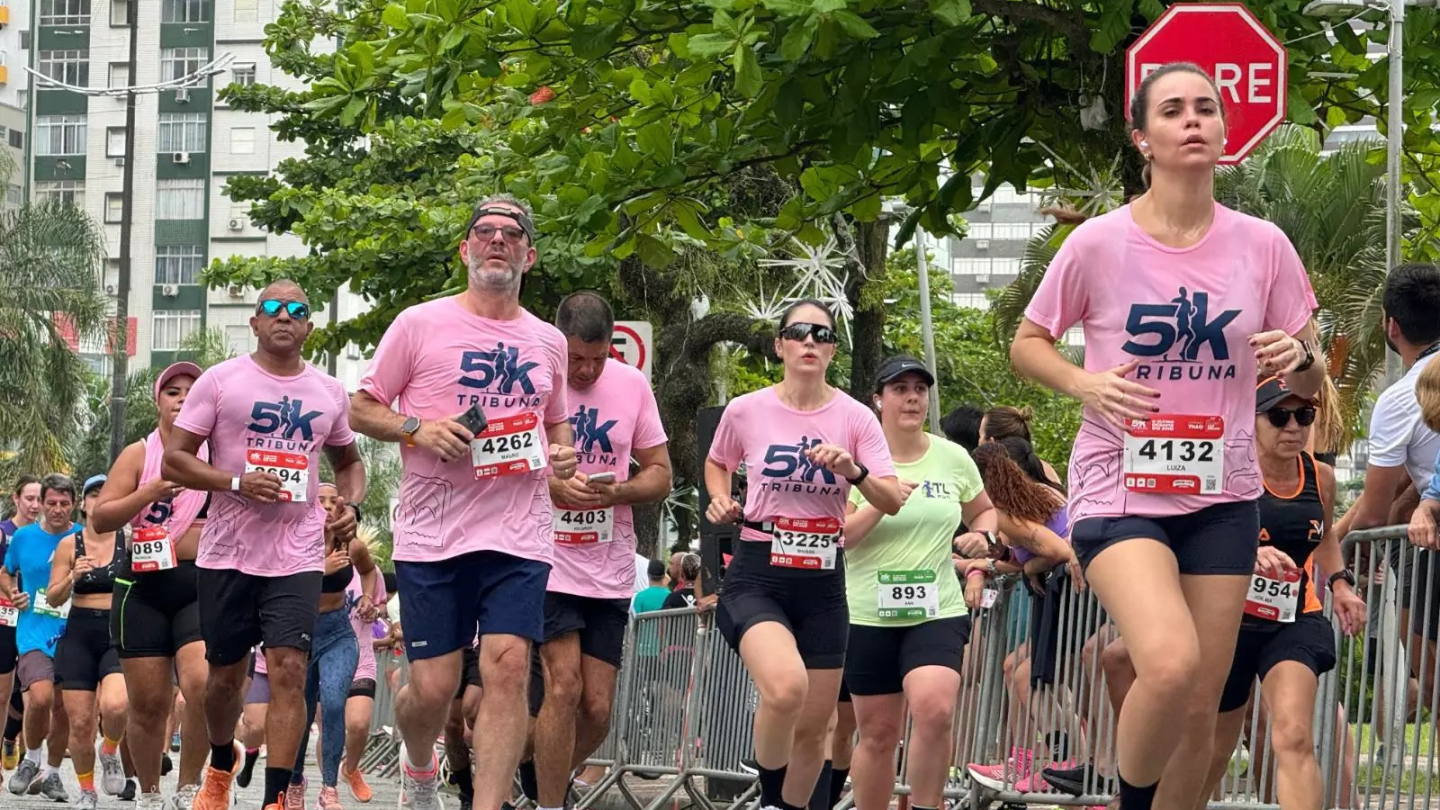 A imagem mostra um grupo de corredores, muitos vestindo camisetas cor-de-rosa com a inscrição "5K Tribuna", participando de uma corrida de rua