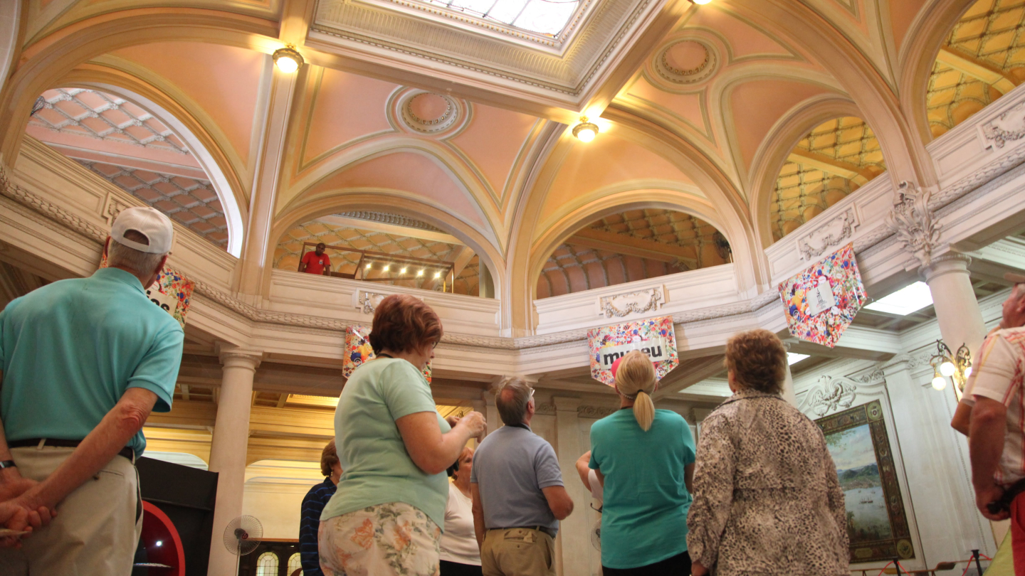 Um grupo de visitantes observa a arquitetura clássica e o teto trabalhado de um museu, caracterizado por arcos elegantes, iluminação suave e um vitral central