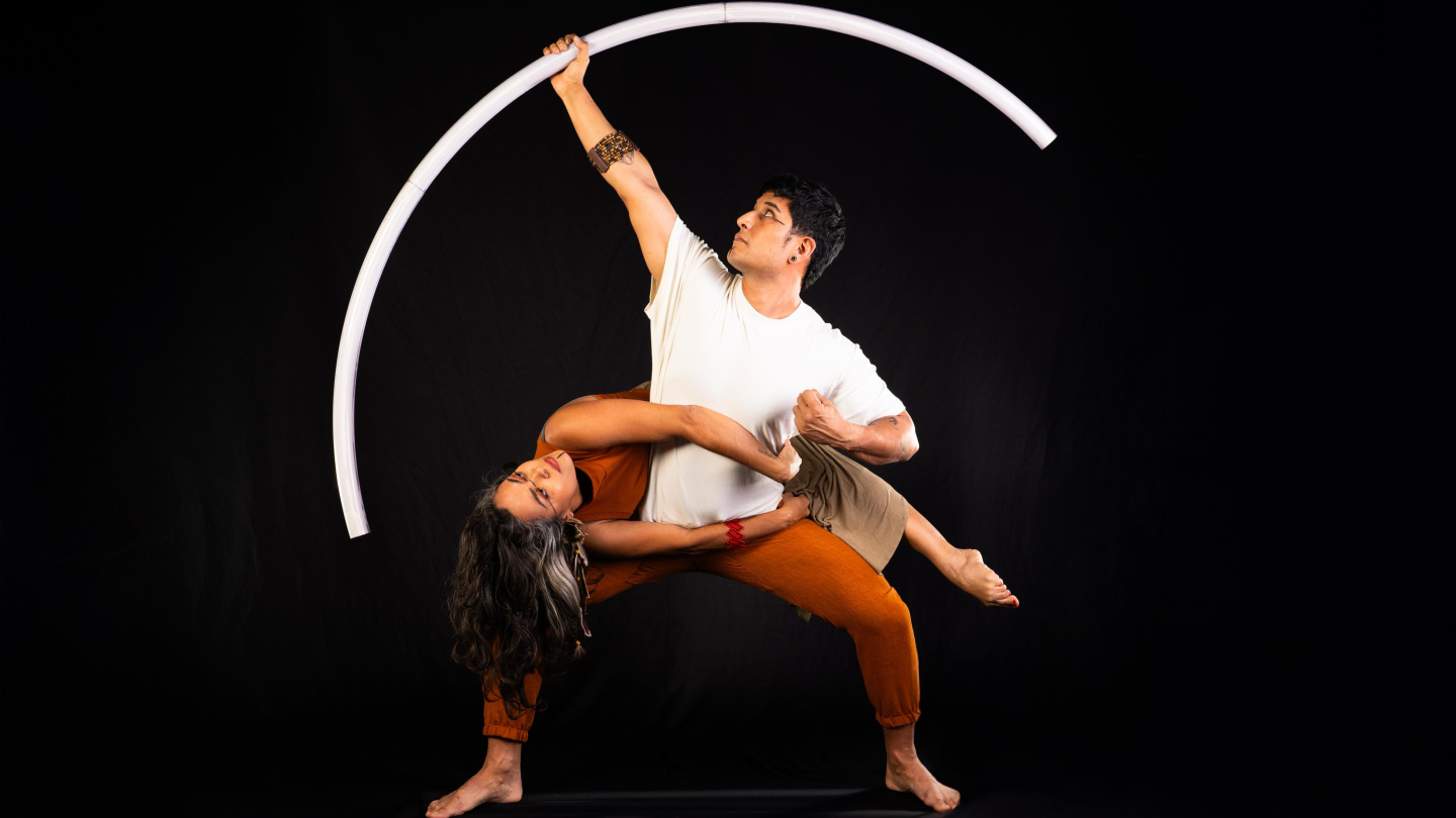 Dois performers realizam uma pose acrobática expressiva contra um fundo preto, onde um homem sustenta uma mulher enquanto segura um grande arco branco luminoso que emoldura a cena