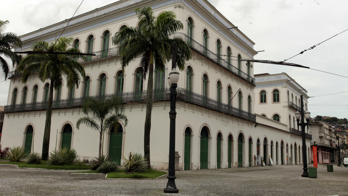 Fachada do Museu Pelé, imponente edifício histórico de arquitetura colonial, com múltiplas janelas verdes e detalhes em azulejos, destaca-se em uma praça arborizada com palmeiras sob um céu nublado