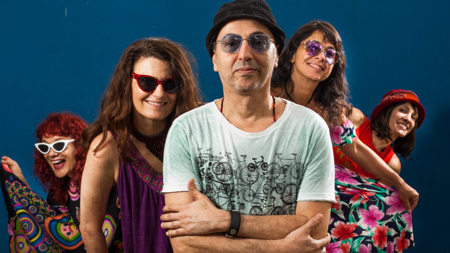 O cantor Zeca Baleiro posa à frente de quatro mulheres sorridentes e vestidas com roupas coloridas, todos usando óculos escuros, contra um fundo azul sólido