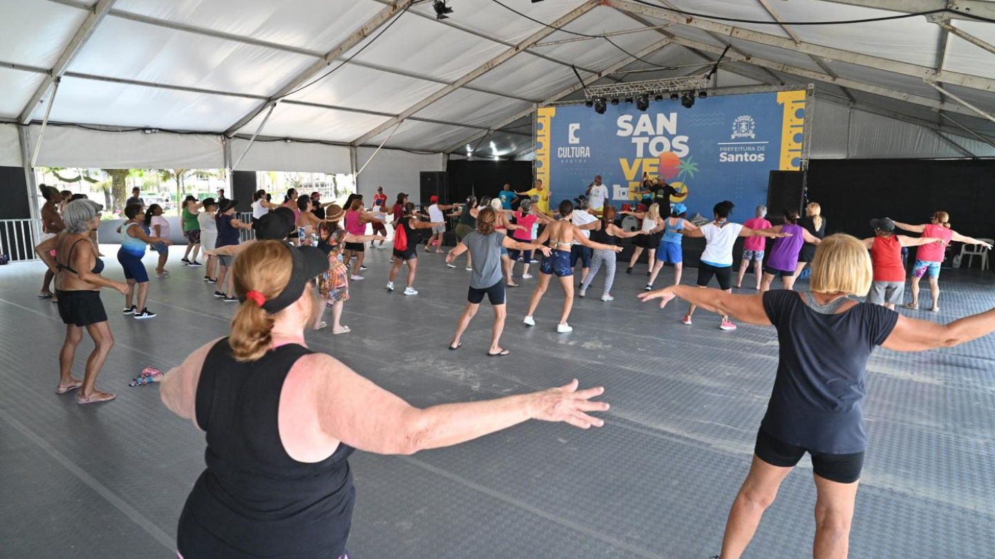 A imagem mostra uma atividade coletiva acontecendo dentro de uma grande tenda montada ao ar livre, com dezenas de pessoas participando de uma aula de dança ou exercício físico. Os participantes, de diferentes idades, estão dispostos em fileiras, com os braços abertos, acompanhando os movimentos orientados por instrutores posicionados à frente do espaço.
