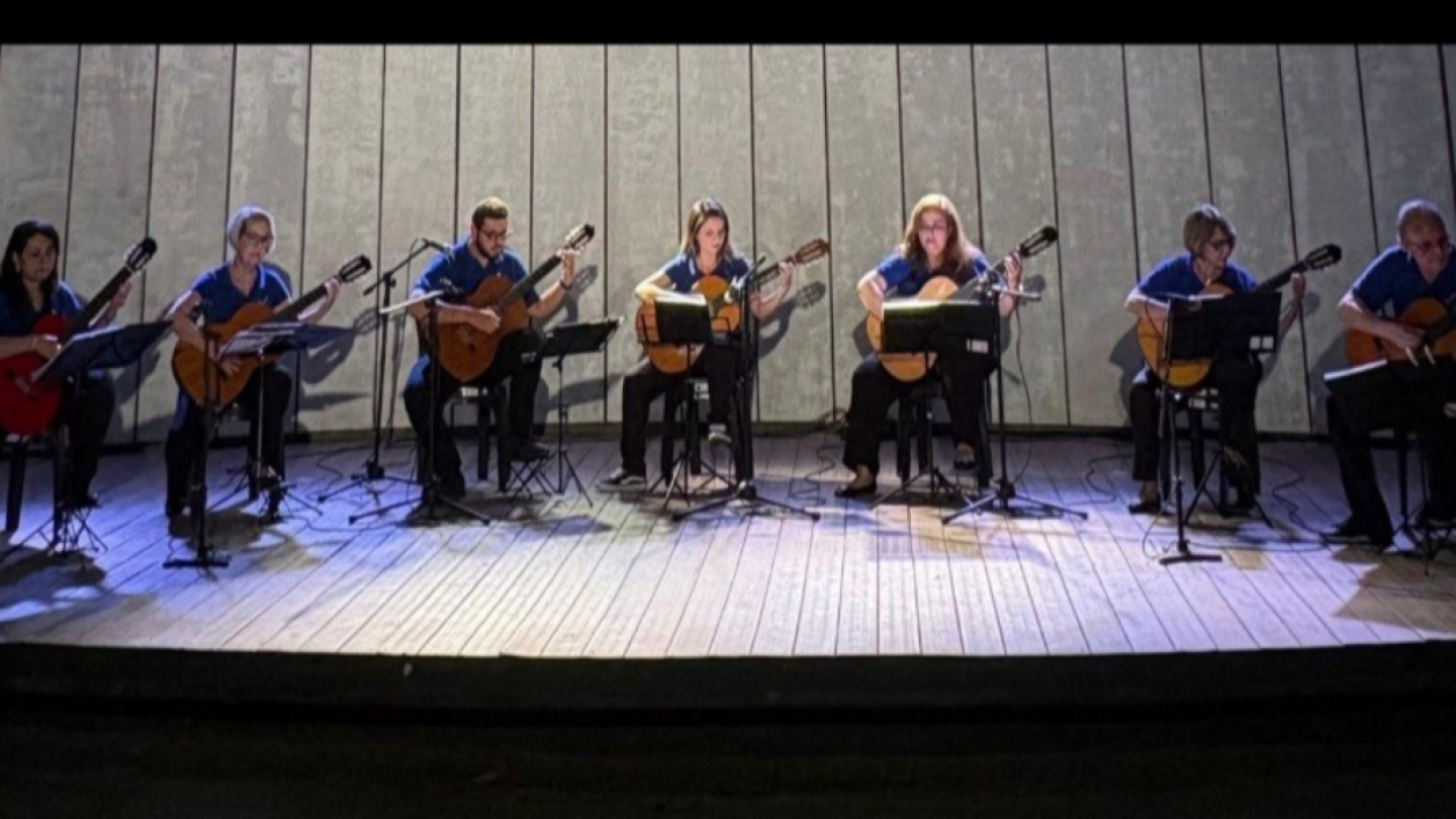 Conjunto musical de sete violonistas (quatro mulheres e três homens) apresentando-se em um palco de madeira