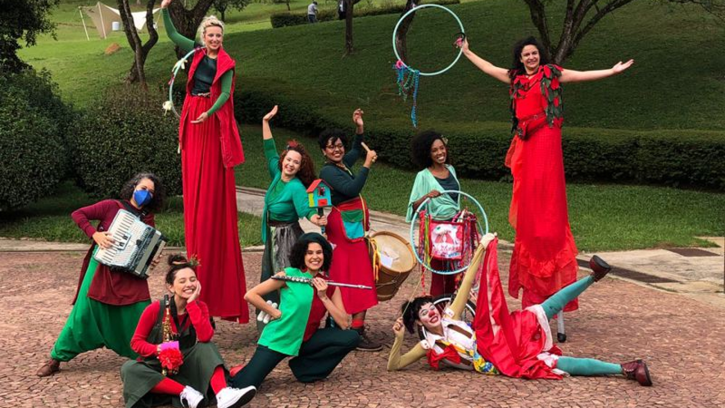 Um grupo circense e musical diversificado posa alegremente em um parque, destacando artistas em pernas de pau, músicos com acordeão e flauta, e uma palhaça em uma performance vibrante