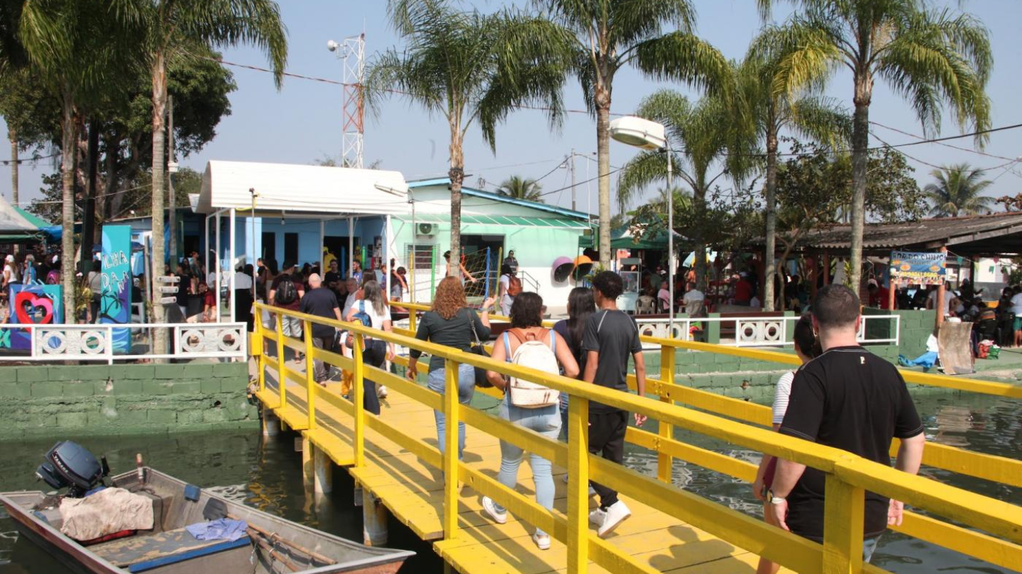 Visitantes atravessam uma ponte amarela de madeira em direção à Ilha Diana, onde casas coloridas e palmeiras compõem o cenário ensolarado à beira-mar
