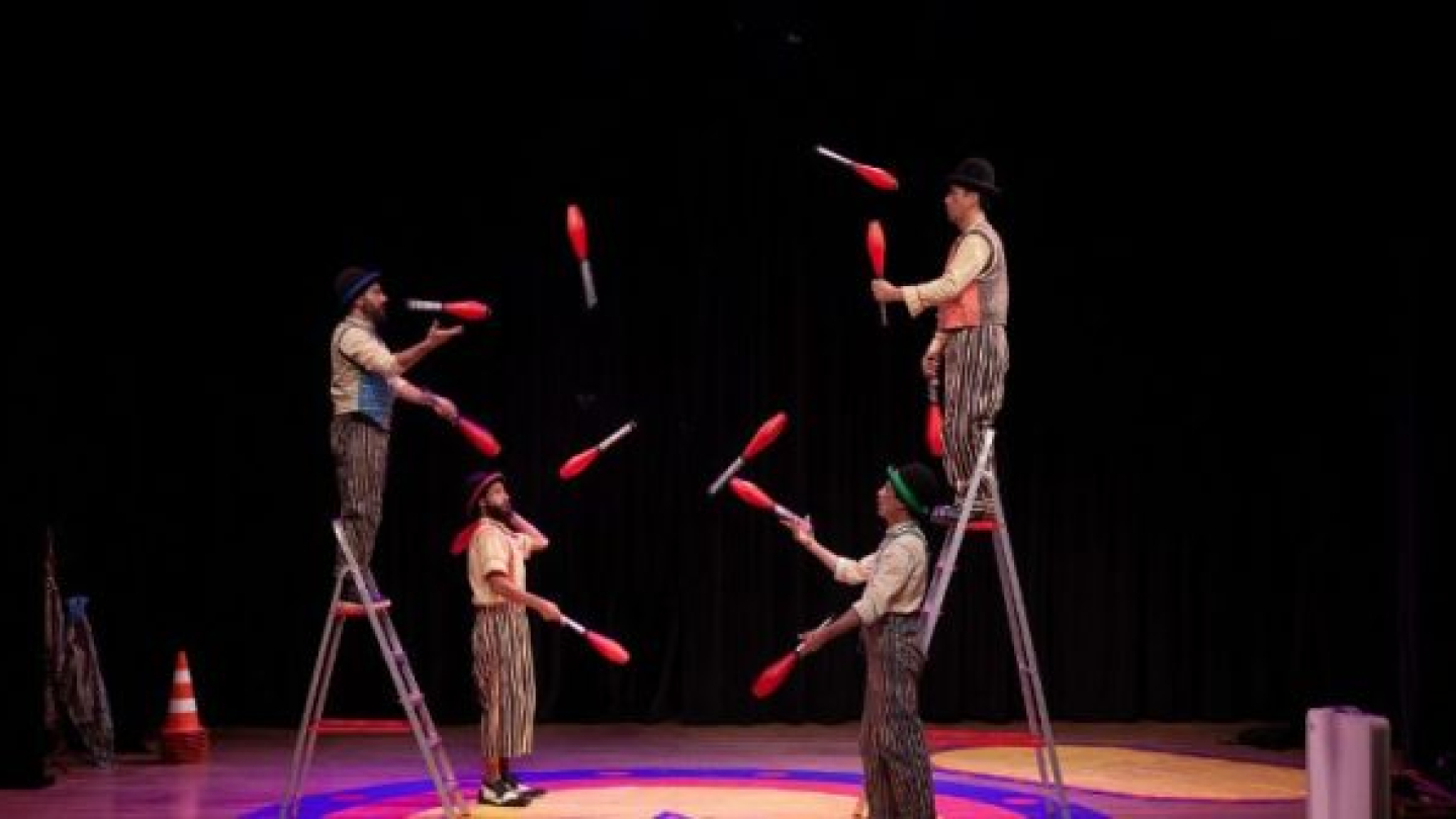 Quatro malabaristas realizam uma performance com pinos vermelhos e brancos em um palco, sendo que dois deles estão equilibrados no topo de escadas metálicas