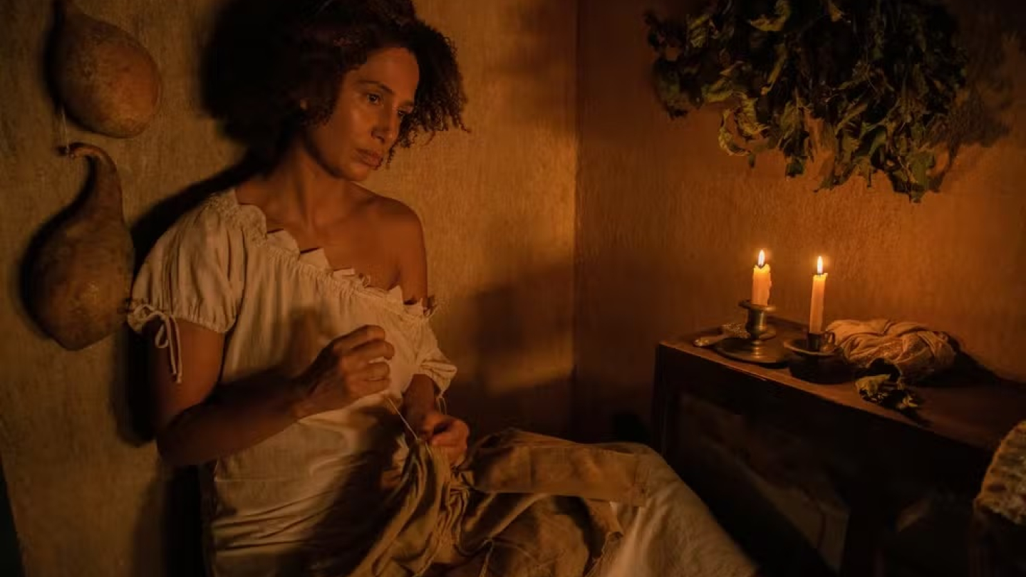 Uma mulher negra costura concentrada sob a luz de velas em um ambiente rústico e sombrio