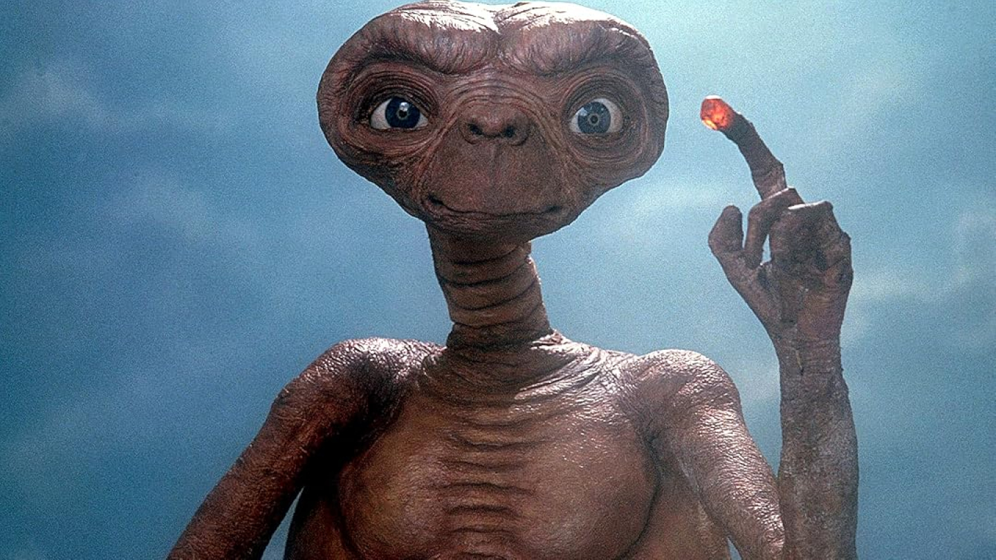O icônico personagem E.T., do filme de Steven Spielberg, aparece centralizado contra um fundo azul claro, apontando para cima com seu dedo longo e iluminado na ponta por uma luz avermelhada