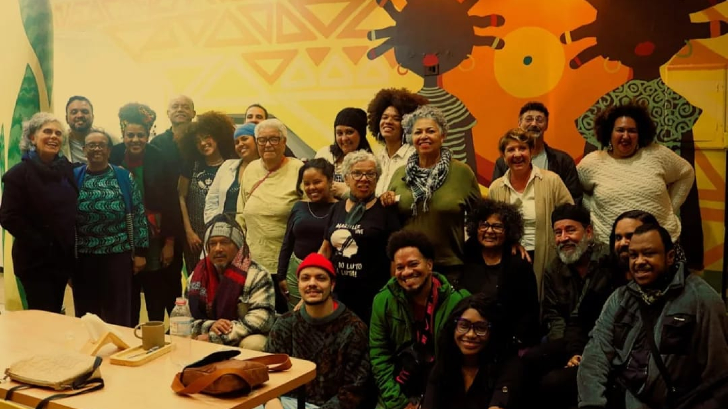 Registro fotográfico de um grupo diverso de pessoas reunidas em um ambiente interno decorado com um mural artístico colorido ao fundo