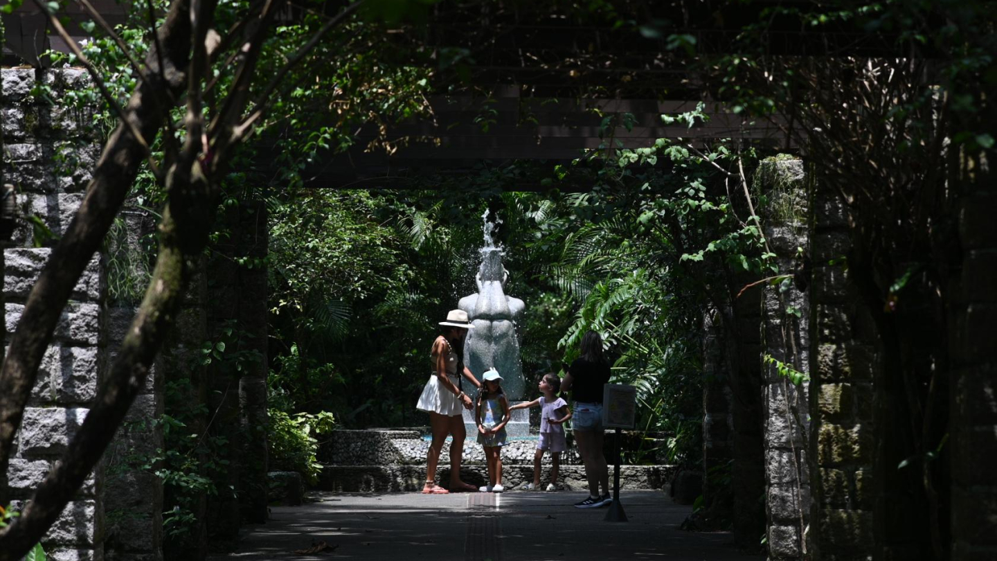 A imagem retrata um momento tranquilo e acolhedor em meio à natureza, em um jardim sombreado e bem preservado. O caminho de pedra conduz o olhar até uma pequena fonte ao fundo, de onde a água cai suavemente, criando um ponto de frescor e contemplação. Arcos formados por árvores e vegetação densa emolduram a cena, filtrando a luz do sol e criando um jogo delicado de luz e sombra.