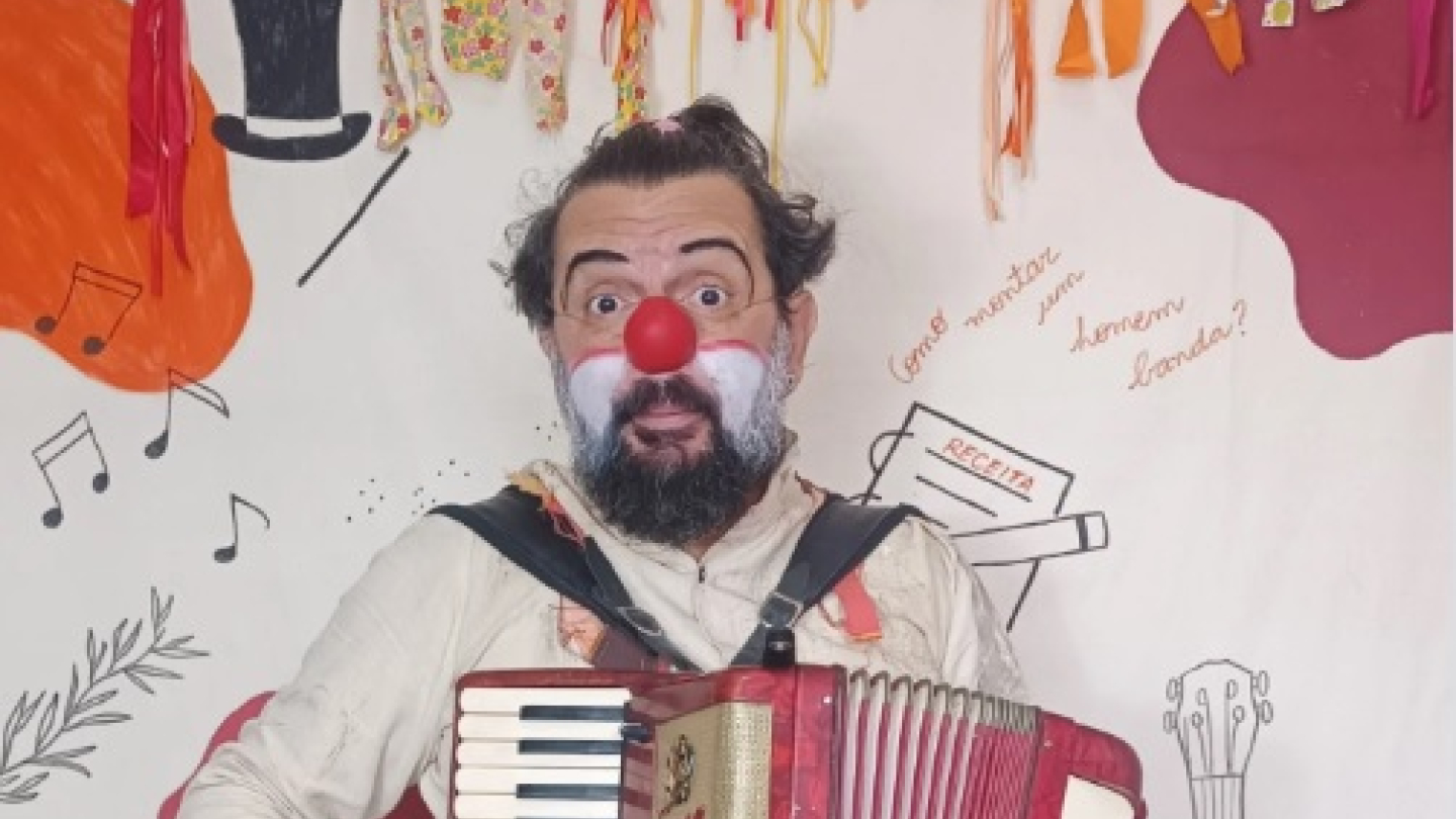 Imagem promocional do espetáculo "Receita de Homem Banda", apresentando um artista caracterizado como palhaço tocando uma sanfona em frente a um cenário lúdico e ilustrado