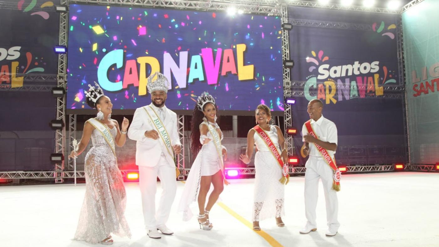 A Corte Real do Carnaval de Santos, incluindo o Rei Momo e a Rainha, celebra alegremente no palco sob um painel luminoso colorido