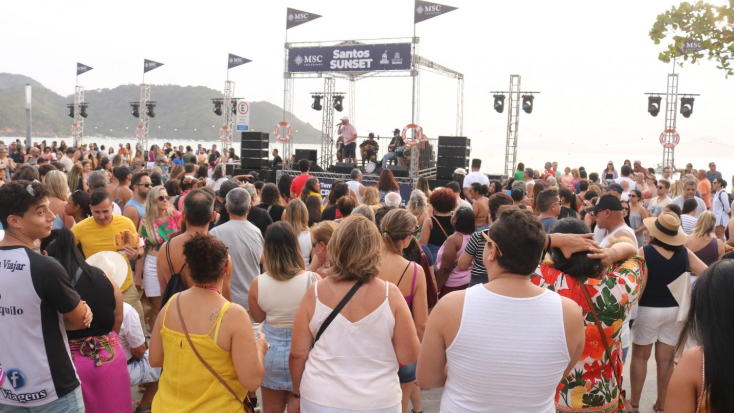 A imagem registra um grande evento ao ar livre na orla, durante o fim de tarde, com um palco montado de frente para o mar. No centro, a estrutura do Santos Sunset se destaca, com banda se apresentando ao vivo e equipamentos de som e iluminação distribuídos ao longo da arena. Ao fundo, o contorno dos morros e o céu claro reforçam o clima de verão e de pôr do sol à beira-mar.