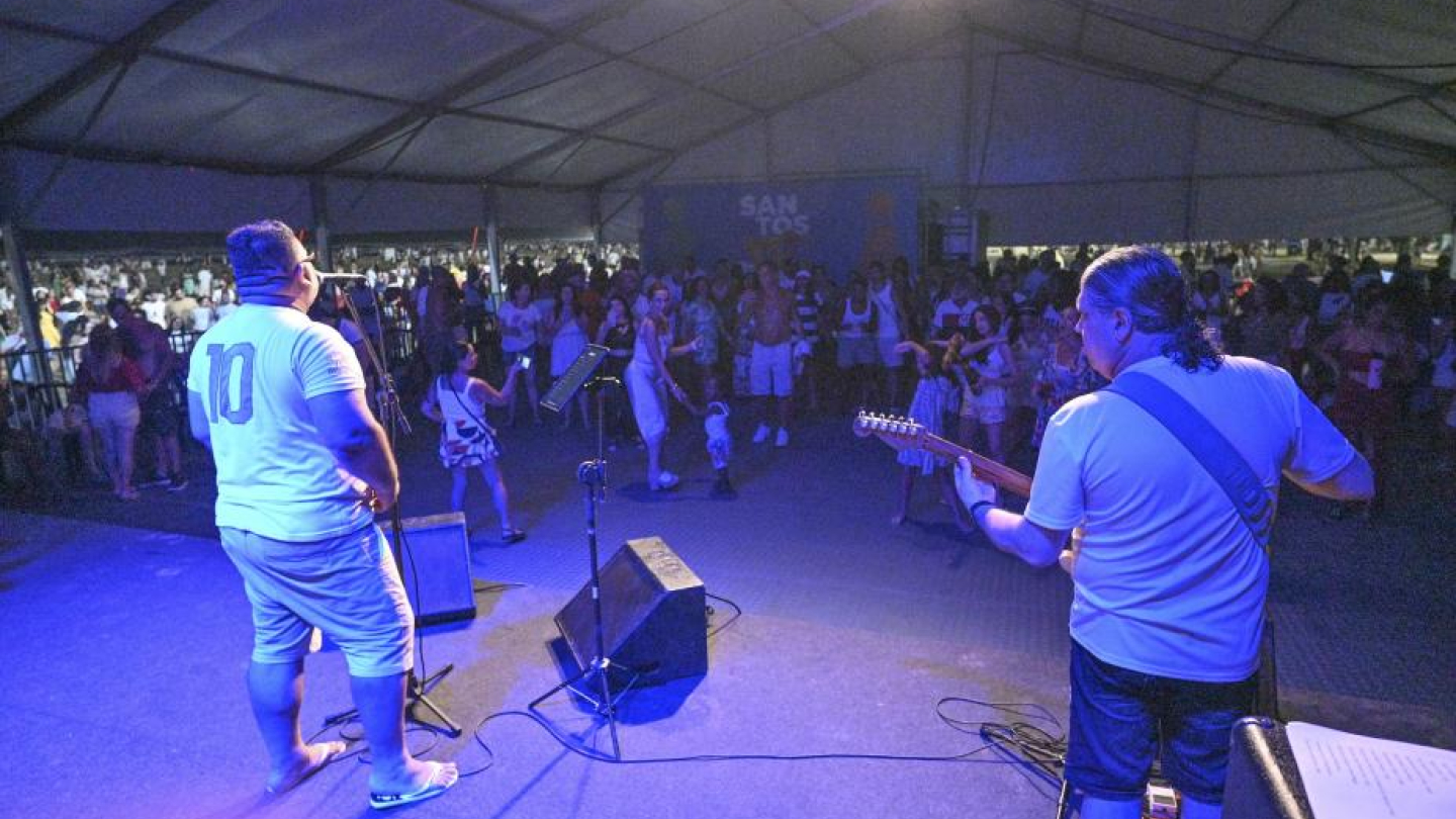 A imagem retrata um show ao vivo acontecendo dentro de uma grande tenda, já no período da noite. A cena é vista a partir do palco, com dois músicos em primeiro plano: um cantor ao microfone e um guitarrista, ambos voltados para o público. A iluminação azul cria um clima envolvente e destaca o palco em contraste com a plateia.