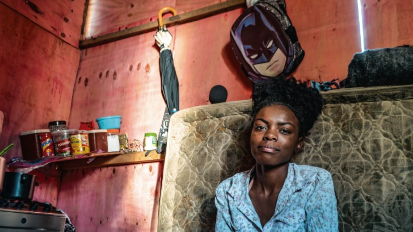 O retrato captura uma jovem mulher negra sentada em um ambiente doméstico humilde, cercada por objetos do cotidiano sob uma iluminação lateral marcante